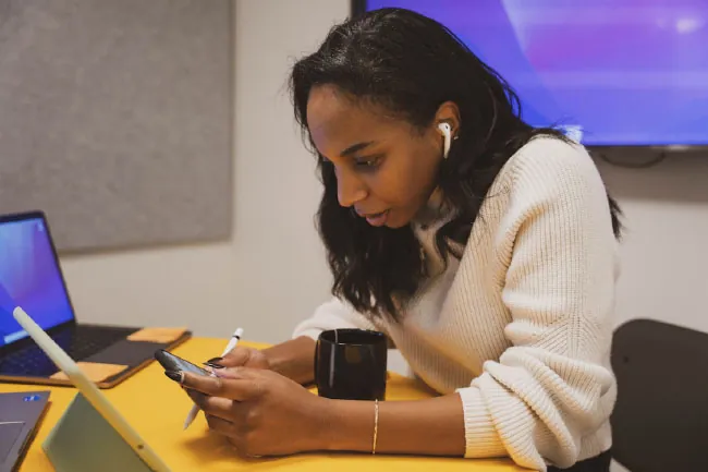 Image of individual sitting in front of a laptop and iPad while looking at their phone, with earbuds in their ears.