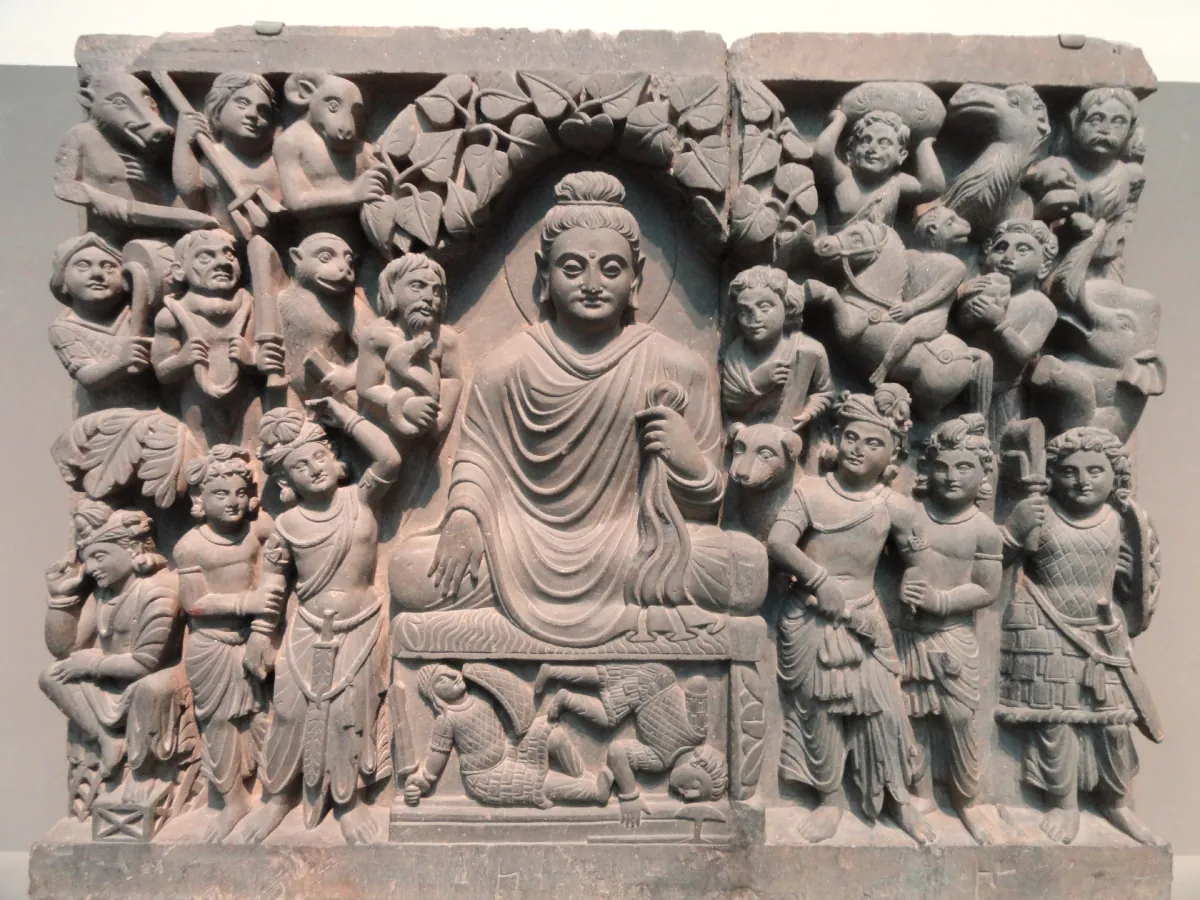 Photo of stone carving of Buddha under an archway. He is surrounded with other carved people and animals, sitting atop a box with dead soldiers inside.