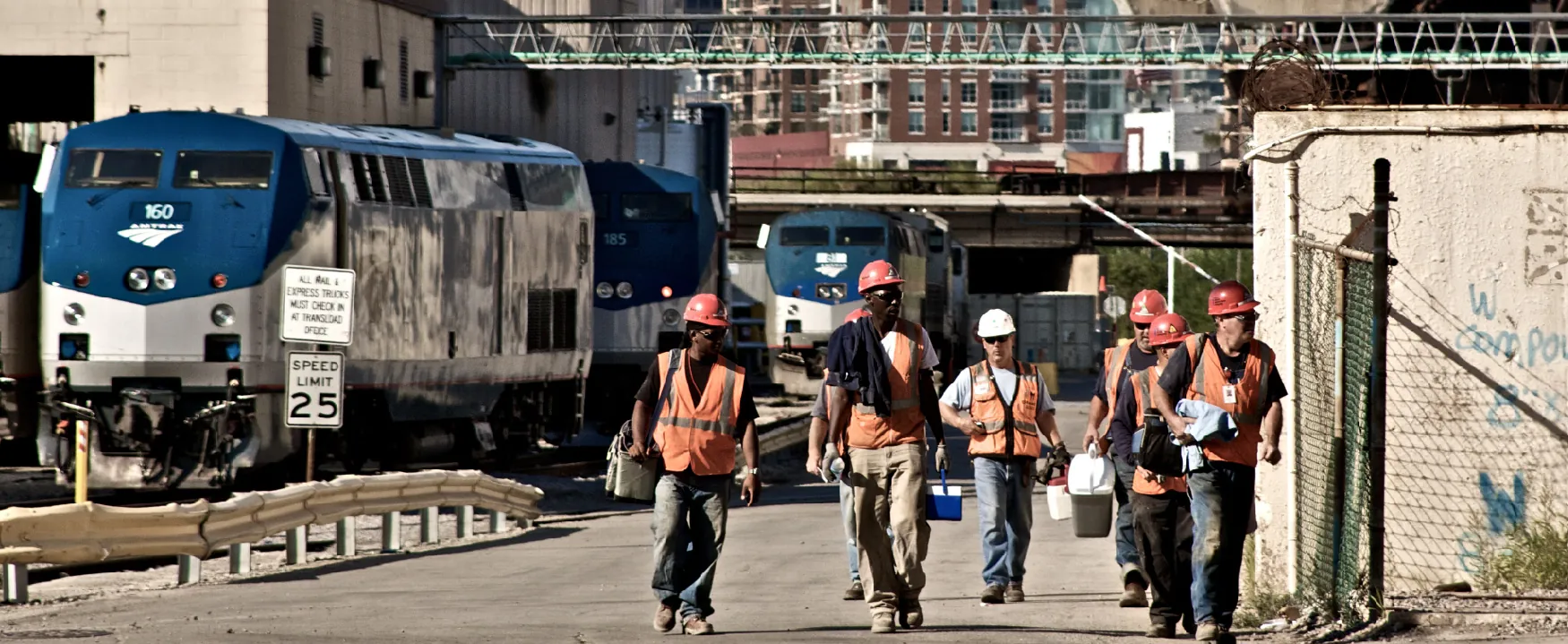 A group of construction workers in safety vests and hard hats walks past parked Amtrak trains in an industrial train yard under a bright sky, with urban buildings visible in the background.