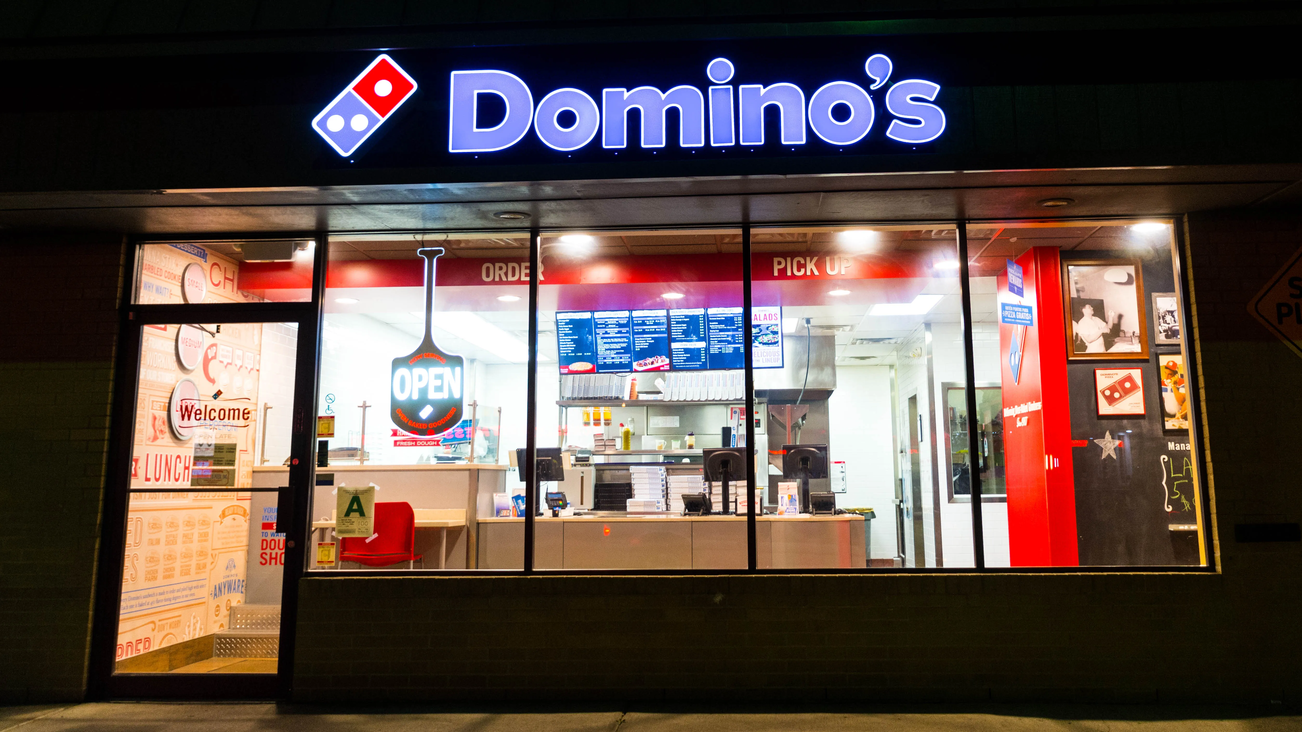 A brightly lit Domino's pizza store at night, its neon sign glowing blue and red. Through the large front windows, the clean interior is visible, featuring digital menus, a counter, and an 'OPEN' sign shaped like a pizza peel.
