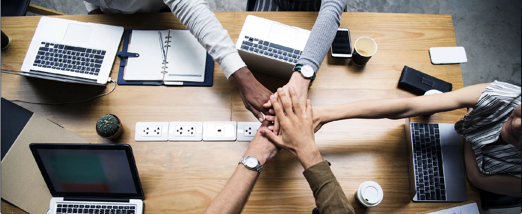 A top-down view shows a diverse team stacking their hands in unity on a wooden table, surrounded by laptops and coffee cups, embodying strong collaboration and shared goals.
