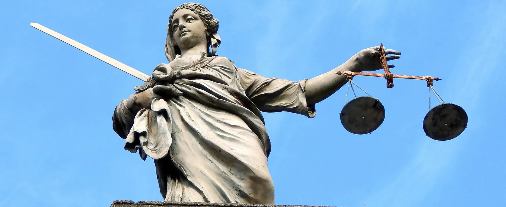 A picture of Arend Ode’s statue depicting Lady Justice. The statue of a woman in Greco-Roman clothing is holding a set of scales in the elevated right hand, and a sword in the left. The statue is located on top of a former justice building in Rotterdam.