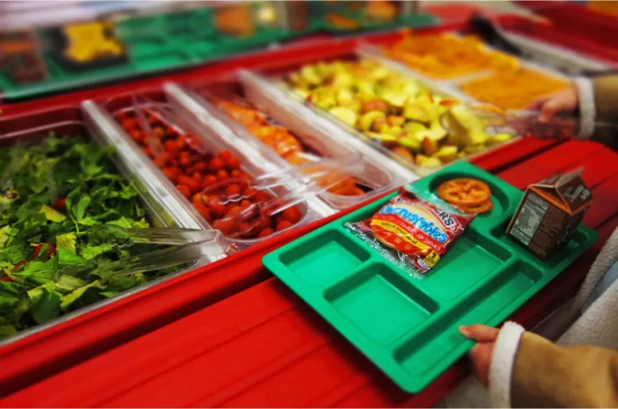 Photo of a salad bar with an individual holding tongs and carrying a tray with an Uncrustables sandwich, tomato slices, and chocolate milk.