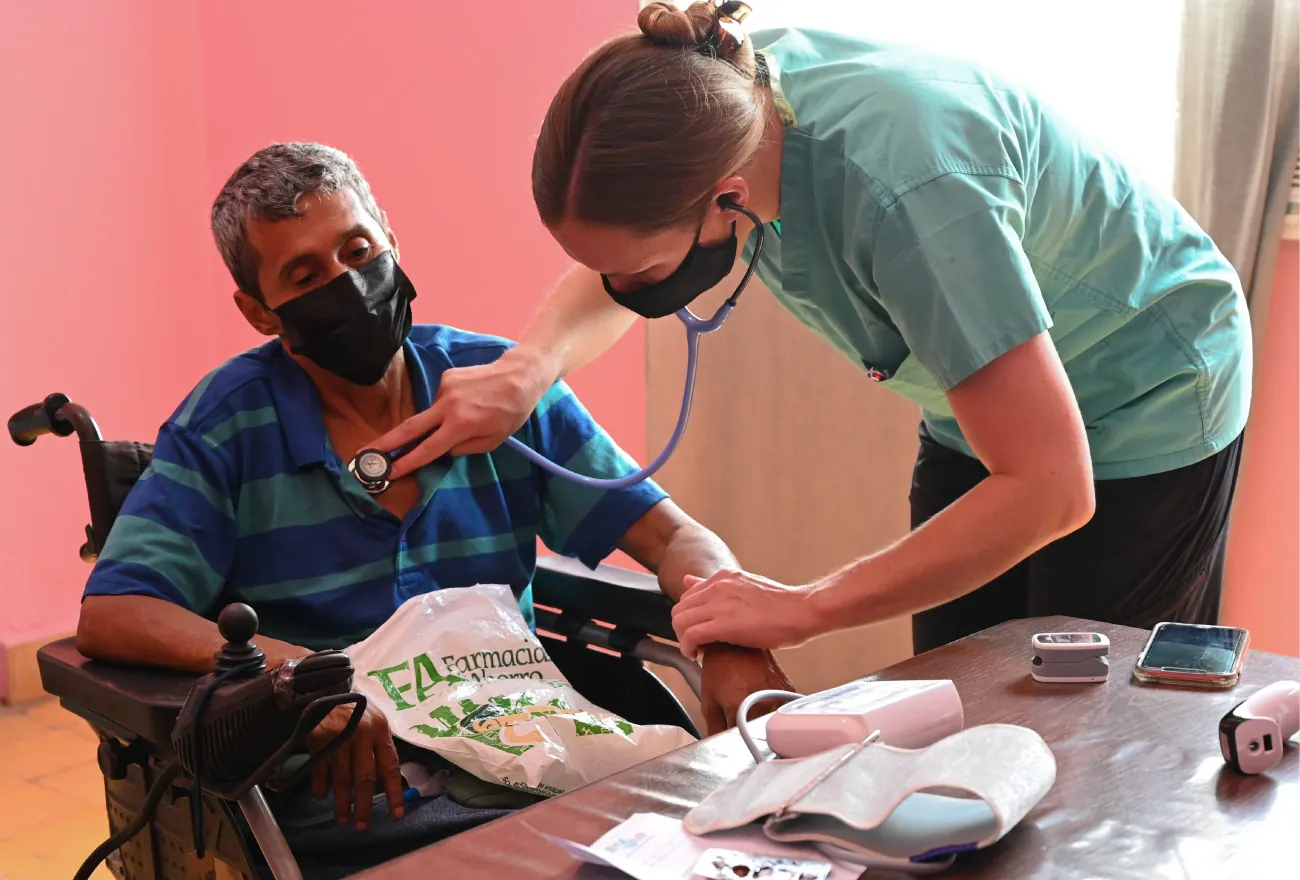 Photo of healthcare worker listening to individual’s chest with stethoscope with other medical equipment on table (blood pressure machine, pulse oximeter, forehead thermometer).