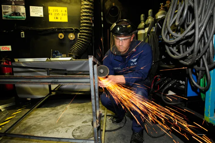 Zdjęcie mężczyzny szlifującego kawałek metalu szlifierką rotacyjną. Dobrze widoczne są iskry wylatujące z tarczy szlifierskiej.