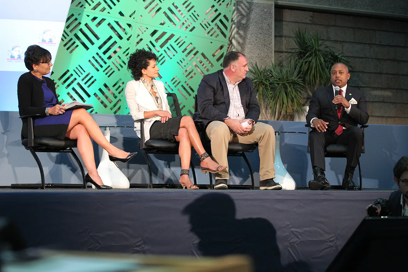A photograph shows Daymond John sitting in a chair on stage, speaking into a microphone.
