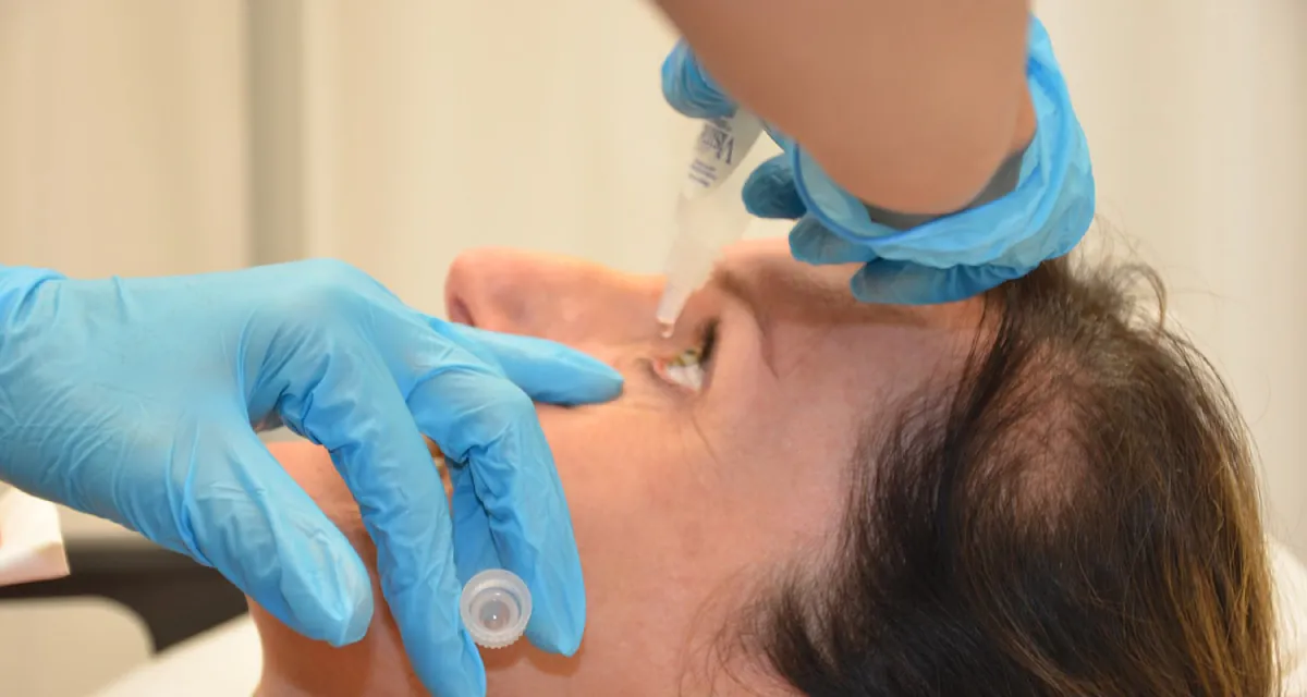 Photo of gloved person holding a person’s eye open along the lower lid and dropping eye drops into their eye.
