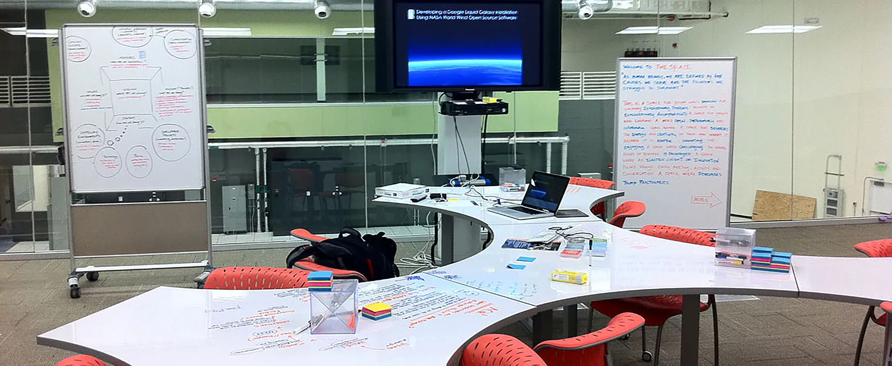 Photo of a classroom with moveable, whiteboard desks, chairs, standing whiteboards, a television, computers, and paper and markers.