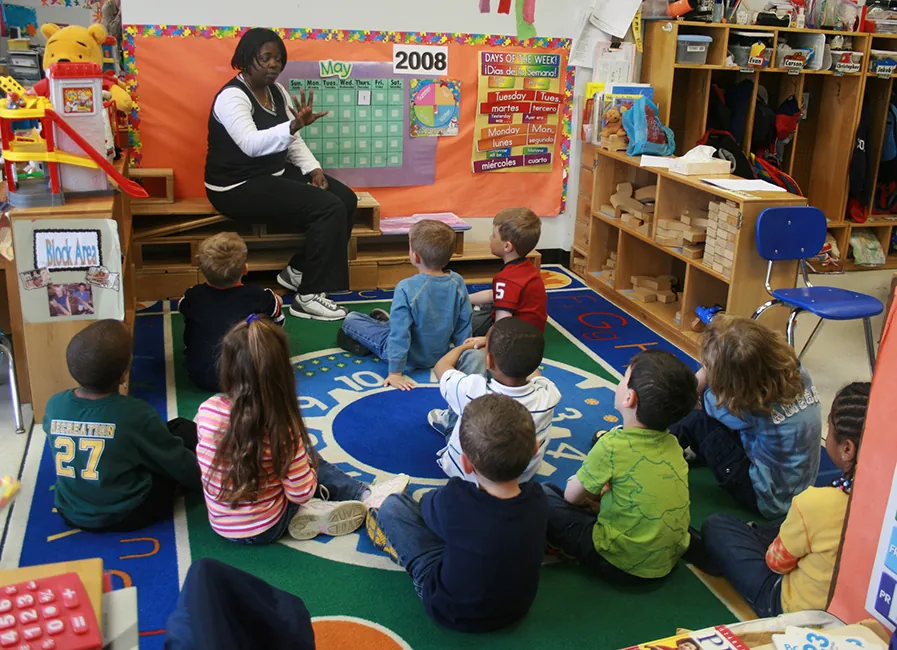 A rear view of a student raising her hand as she sits at her desk. A teacher standing at the front of the classroom is pointing at the student.