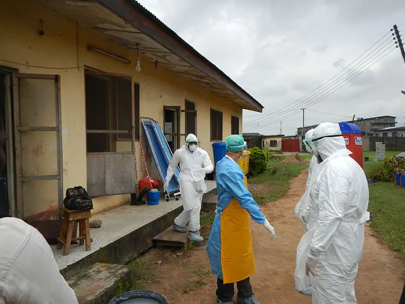 Several people dressed in personal protective equipment outside of a one-story stucco building. Their gear includes face masks, gloves, foot coverings, and goggles.