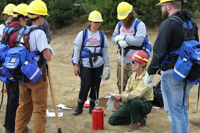 Photograph shows several people wearing hard hats and Team Rubicon shirts, with outdoor gear and shovels, gathered around someone kneeling with a piece of firefighting equipment.