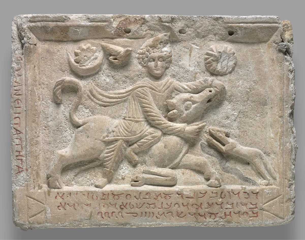 An image of a rectangular pale brown stone is shown on a gray background. The edges of the stone are worn and broken in places. The top edge shows three holes. Letters are engraved along the left edge and scripted images are engraved in red along the bottom in three rows. The middle shows a raised image of a figure in a striped shirt and pants with a long flowing cape sitting atop an animal with four legs, long bushy tail, and a long snout. The figure has round eyes and a large hat on his head. His right arm encircles the head of the animal he sits on and his left hand is close to the nose of the animal. The animal he is sitting on has bent legs and is close to the ground. Another small animal standing up on its hind legs reaches toward the front of the larger animal. A round sun with rays and broken in places is carved in the top right and a crescent moon with a flowery image inside is carved at the top left. A small, winged creature is seen to the left of the figure’s head.