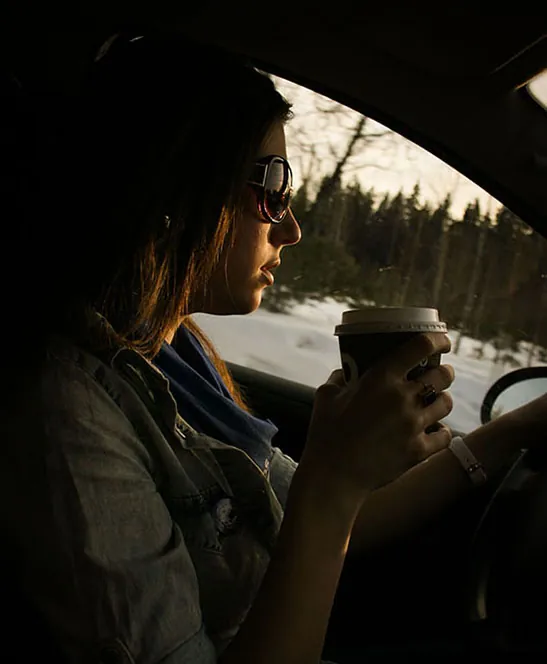 A person wearing sunglasses sits in the driver's seat of a car with one hand on the steering wheel while the other holds a to-go coffee cup.