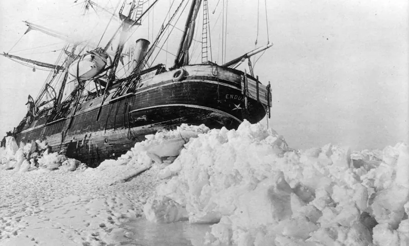 A black and white photograph of a shipwreck, with an old expedition-style sailing ship stuck in ice.