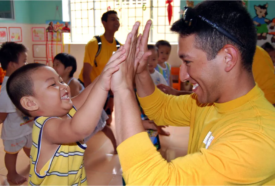 Smiling adult and child high fiving each other with both hands in room full of other children and adults.