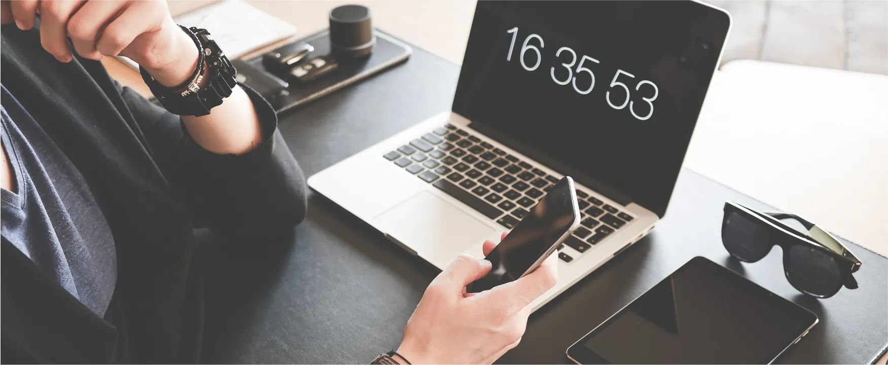 A person sits at a desk holding a cell phone and wearing a watch. A laptop, iPad, glasses, and other devices lay on the desk.