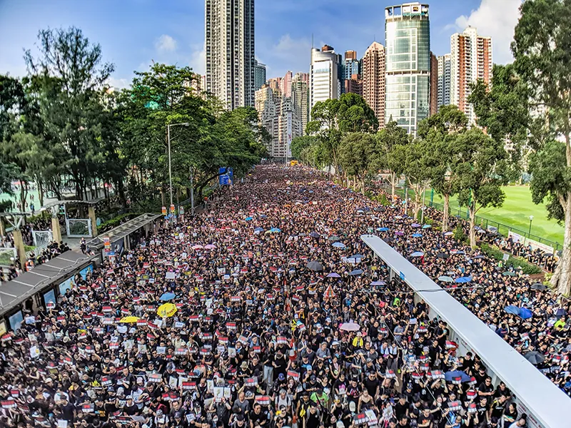 An overhead photograph shows a large, tree-lined, city street completely filled with people. Skyscrapers are visible in the background.