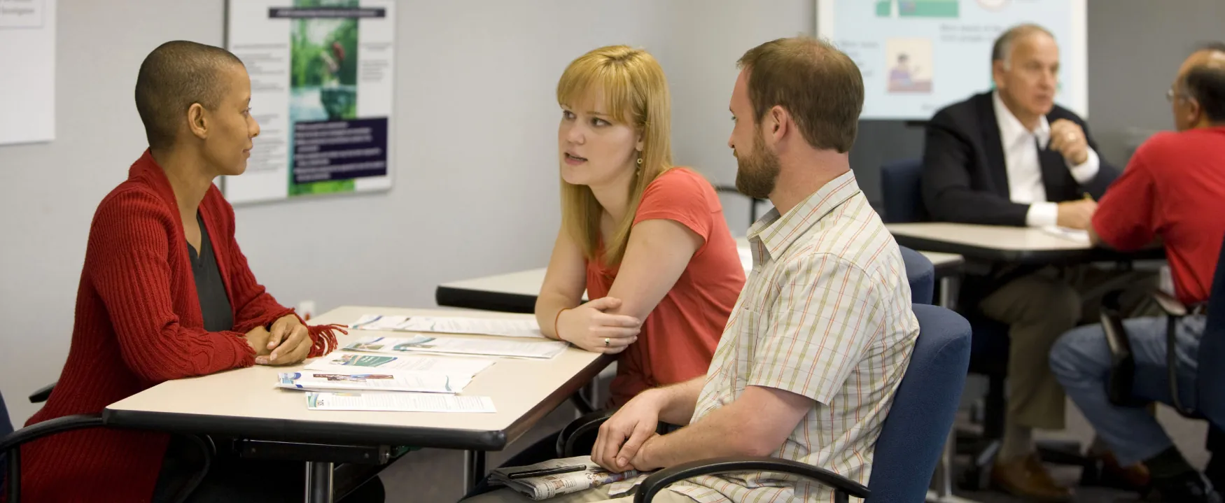 In the foreground, a man and a woman on one side of a table speak to another woman on the opposite side.  In the background, some other groups speak at tables.  