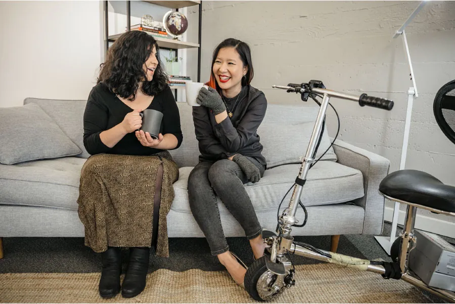 Photo of two individuals sitting on a couch talking, with a mobility scooter parked next to them.