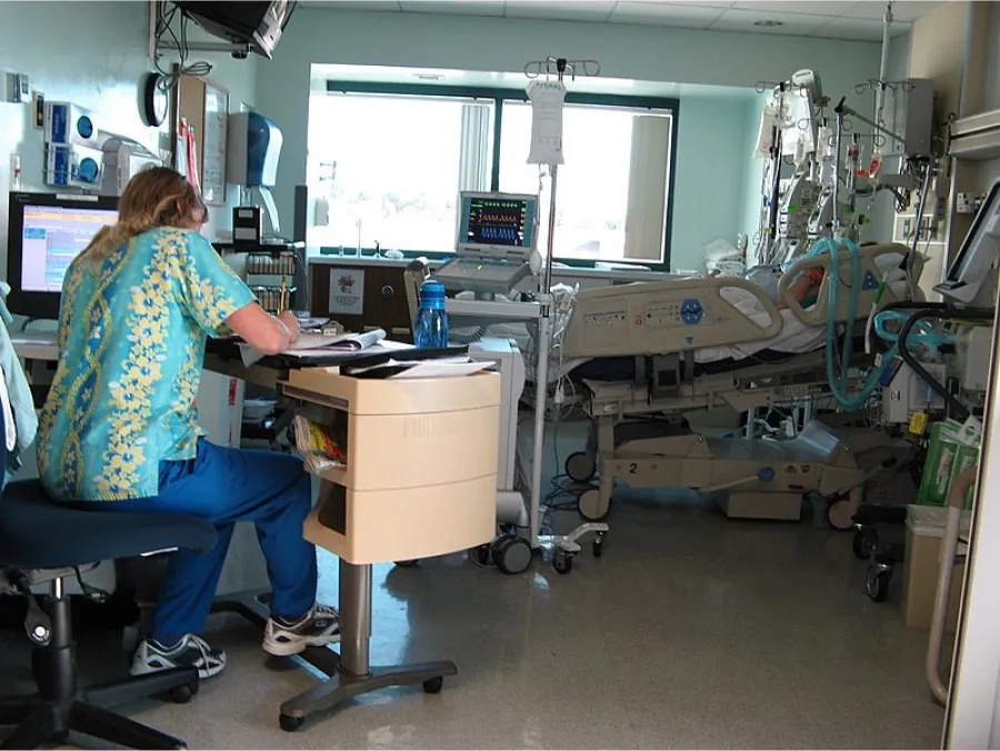 Photo of healthcare worker sitting at a desk in a hospital room with an individual lying in a bed surrounded by machines.