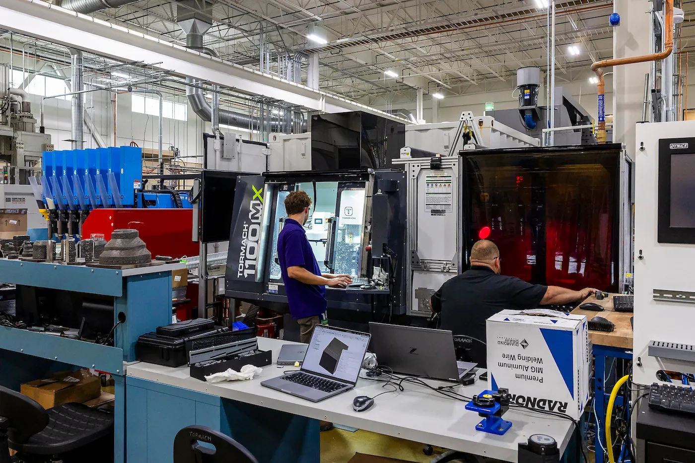 Two people view two different types of machines in a manufacturing facility. They are surrounded by additional machines as well as a desk with a laptop displaying a design. 