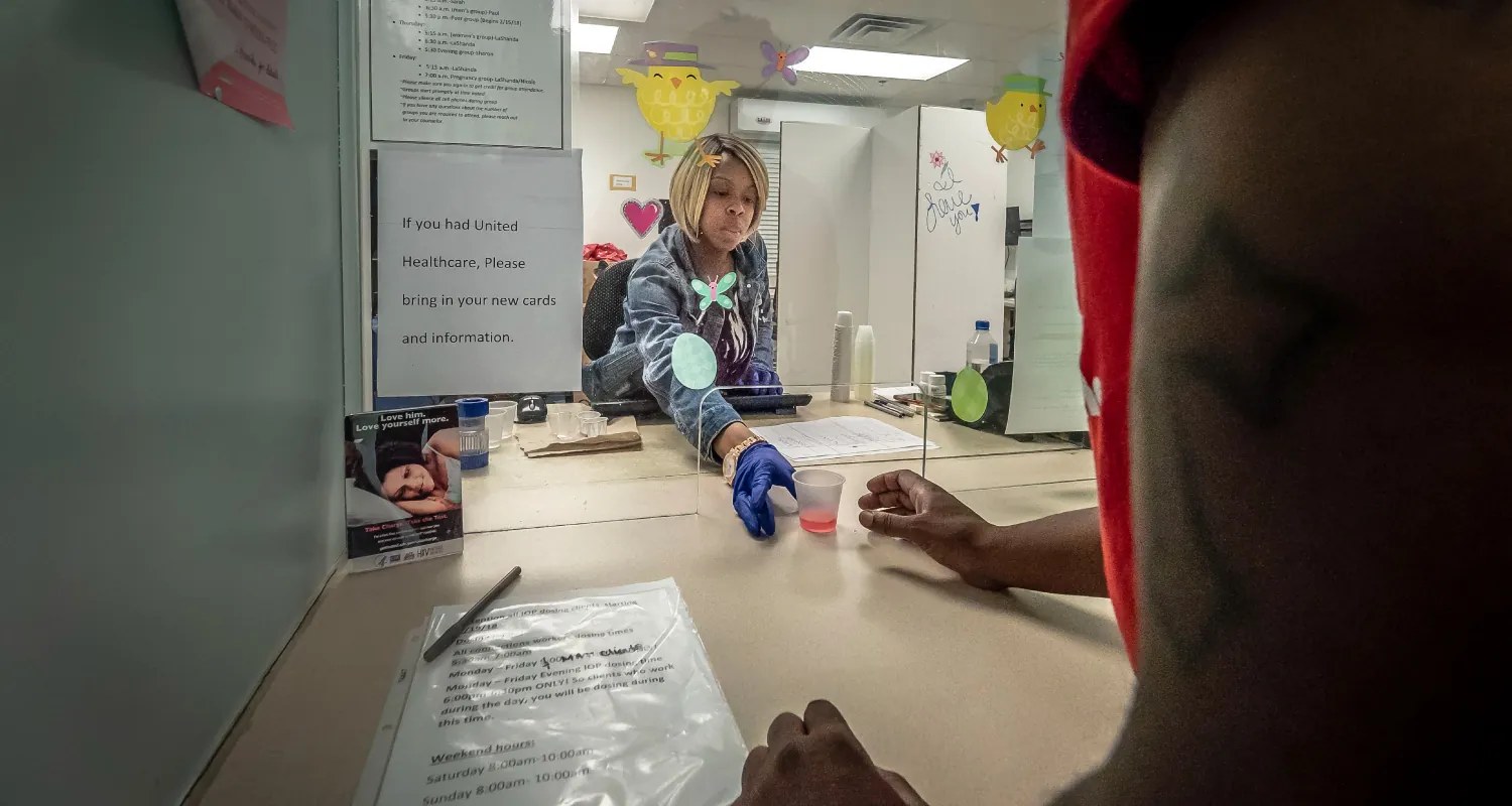 A nurse sitting at a desk, wearing latex gloves, passes a small plastic cup containing a small amount of fluid to a client on the other side of a clear plastic partition.