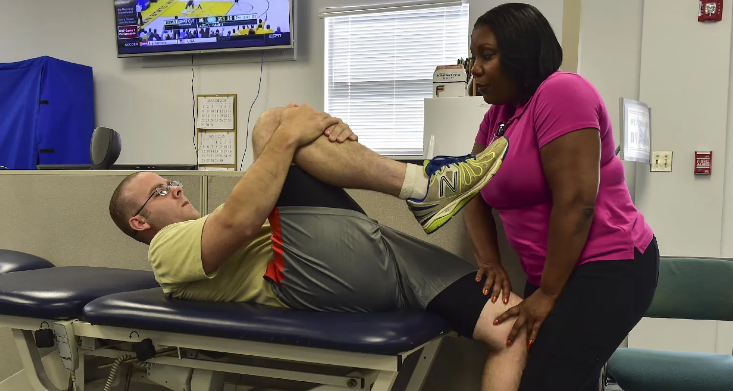A man is doing a leg stretch on a treatment table with assistance from a woman in a physical therapy clinic, while a sports channel plays on a TV in the background.