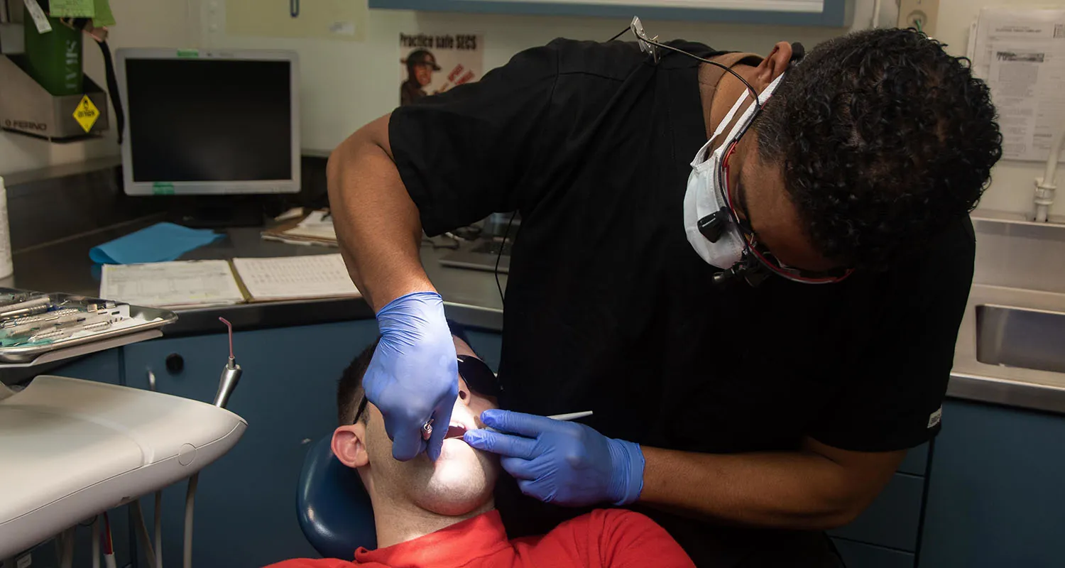 A soldier in uniform lies on a table. A dentist sits beside them, wearing a face mask and glasses with magnifying lenses and a light. In the background, an assistant holds papers and works on a computer.