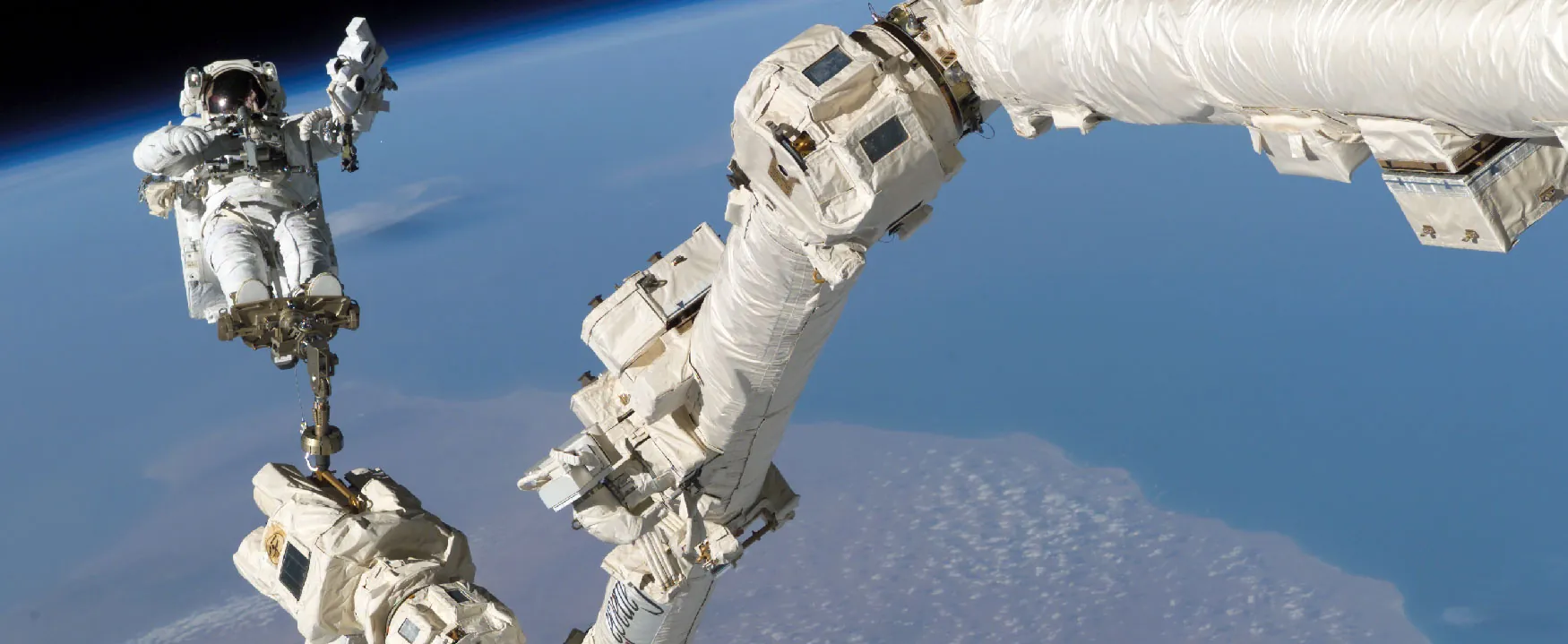 An astronaut on a spacewalk, tethered to a robotic arm, with the stunning view of Earth's atmosphere and surface below.