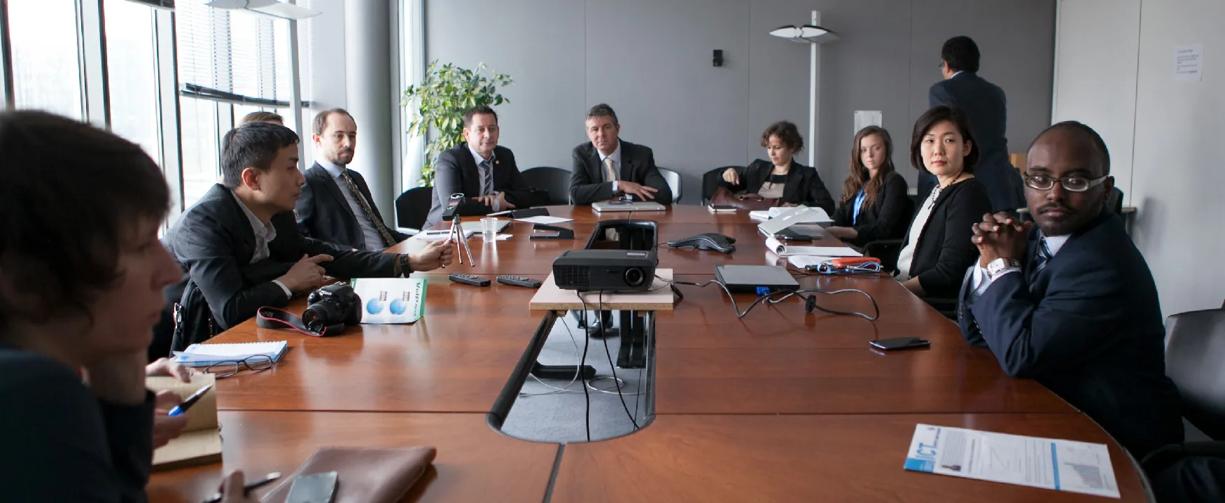 A diverse group of professionals attend a business meeting around a long wooden conference table with a projector and other tech. Some look at the camera, others are engaged in discussion.