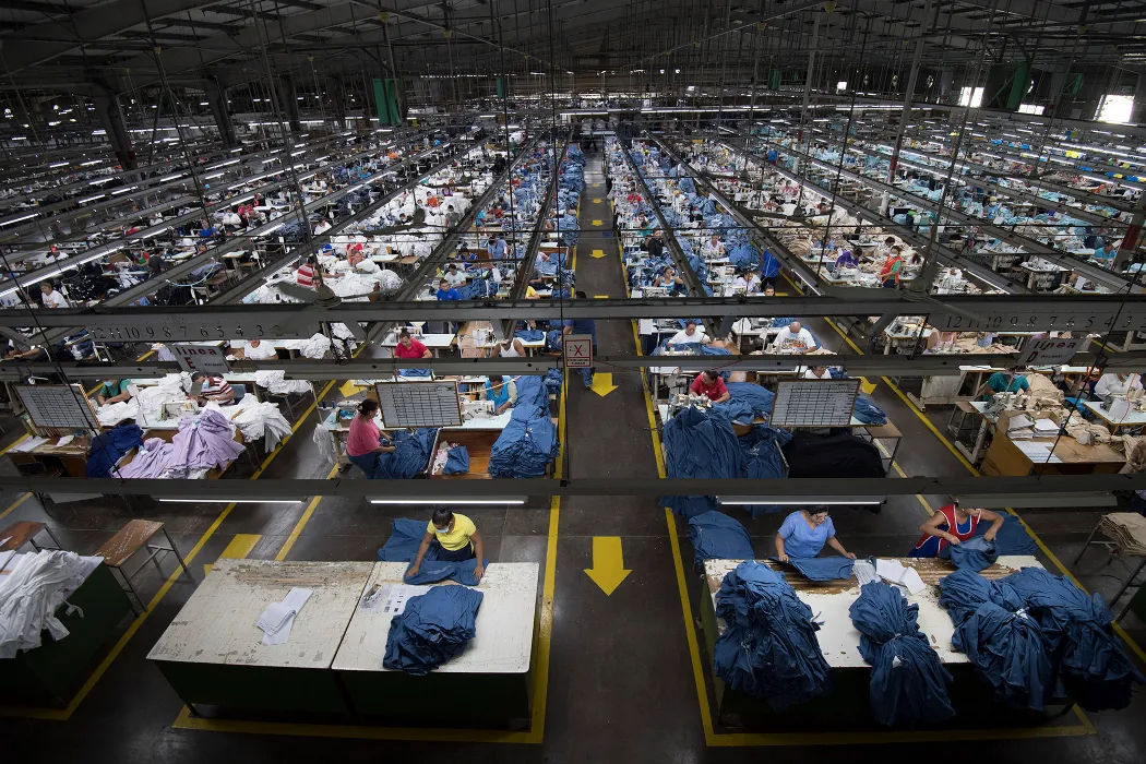 Garment workers work in a vast, warehouse sized room. Large tables are arranged in rows throughout the room, with one or two workers working with fabric on the tables. Arrows are painted on the floor.