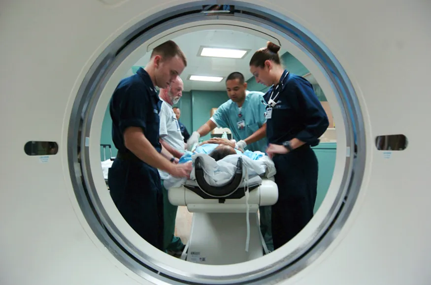 A photographic image taken through the port of a C T scanner, showing a patient on a stretcher surrounded by three nursing staff and a doctor who are taking the patient’s C T scan.