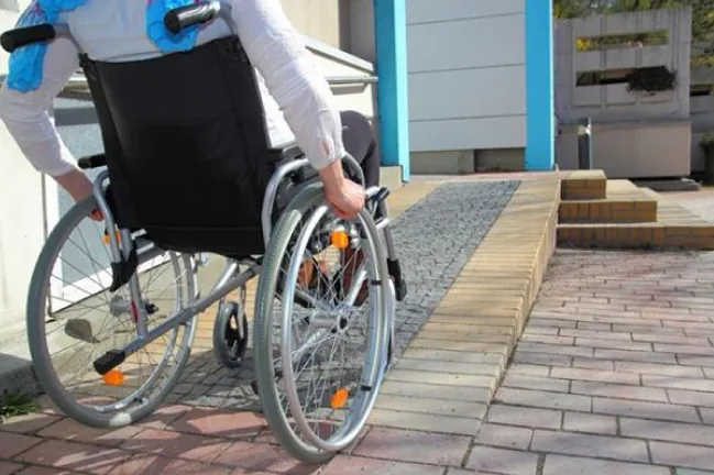 A photograph of a man in a wheelchair rolling up a long ramp leading to an entrance.