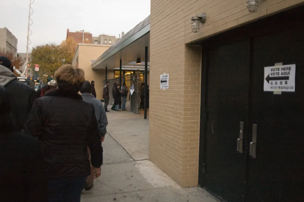 People are shown standing outside a building in line. Signs on the building read “vote here” in various languages.