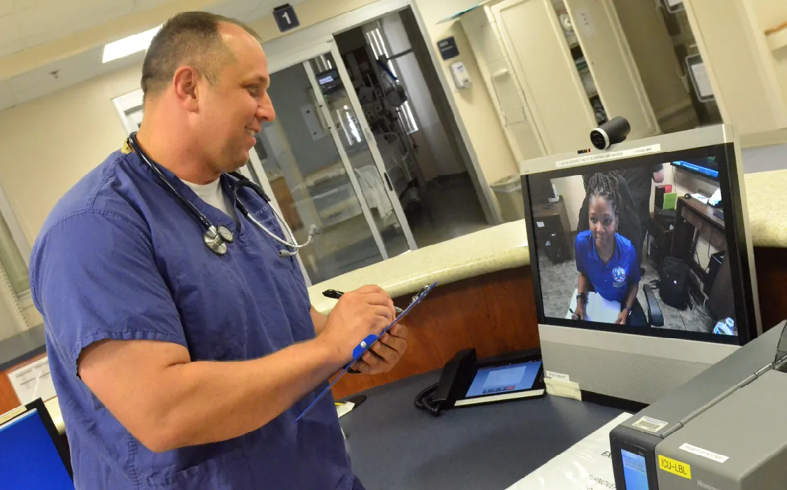 A healthcare provider stands in front of a computer screen and writes on a clipboard. The computer screen shows a person seated in an office.