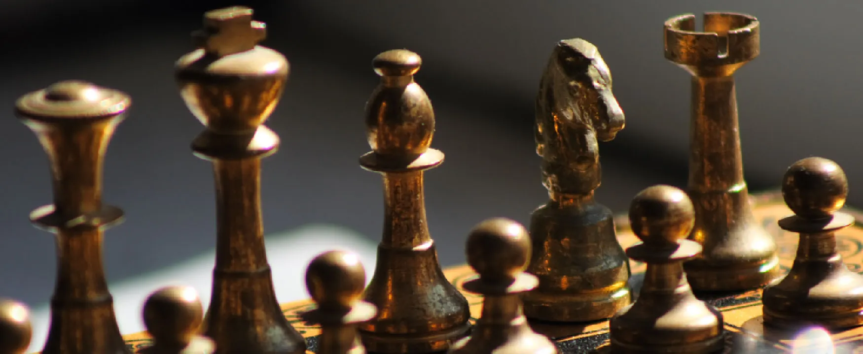 A close-up shot of several gold-colored chess pieces on a chessboard, dramatically lit from the side. The pieces, including a king, bishop, knight, and rook, cast long shadows against a dark background, highlighting their intricate details and metallic sheen. The lighting creates a strong contrast and depth.