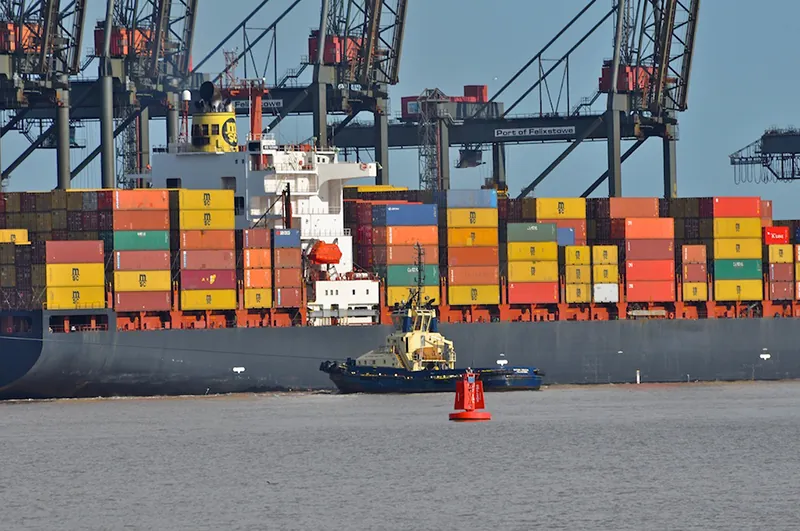 Shipping containers are stacked on top of each other on a barge in the water. A smaller boat is in front of the barge.