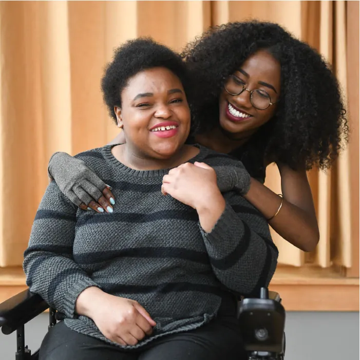Photo of individual sitting with another individual hugging them from behind. Both are smiling.
