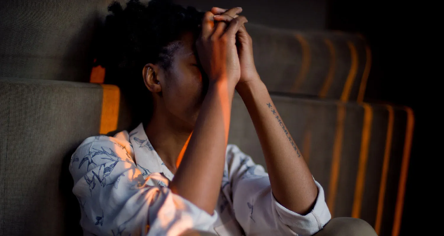 Photo of individual sitting behind a couch with her arms resting on her knees, covering her face.