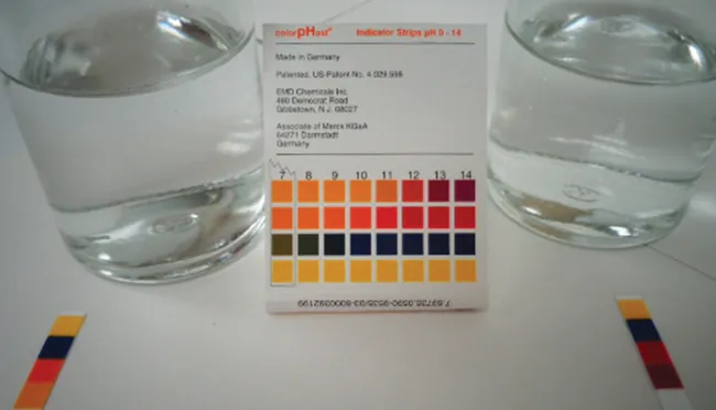 This photo shows two glass containers filled with a transparent liquid. In between the containers is a p H strip indicator guide. There are p H strips placed in front of each glass container. The liquid in the container on the left appears to have a p H of 10 or 11. The liquid in the container on the right appears to have a p H of about 13 or 14.