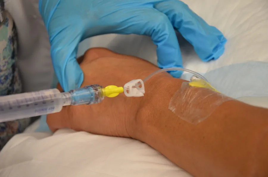Photo of gloved hand holding ungloved hand while a syringe is attached to a tube taped to the ungloved person’s arm.