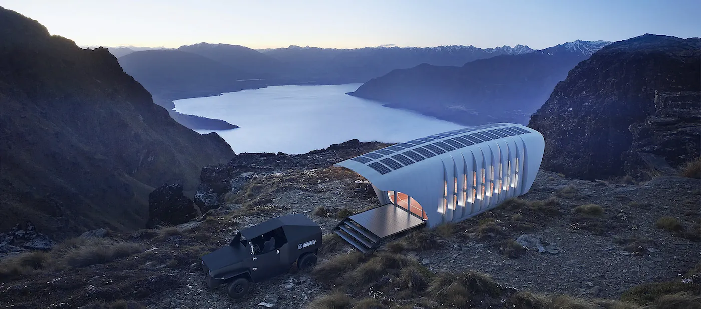 A cylindrical structure with narrow windows along its sides, solar panels on top, and a step opening sits on a rocky bluff above a body of water. A sport utility vehicle is parked in front. 