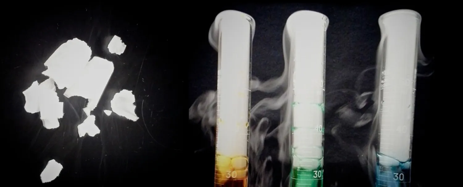 This figure shows pieces of a white substance which appear to be sublimating. To the right of these pieces are three graduated cylinders. Each cylinder holds a different color liquid, and above the liquid, the cylinders are filled with a fog-like substance. This fog-like substance swirls out of the top and around the outside of the cylinders.