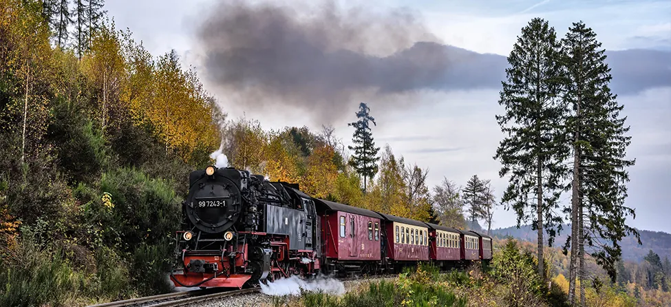 A steam engine and several passenger cars are shown traveling down a train track. The train has some people on board.