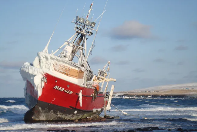 Photograph shows a commercial fishing vessel grounded in shallow water and partially covered in ice.