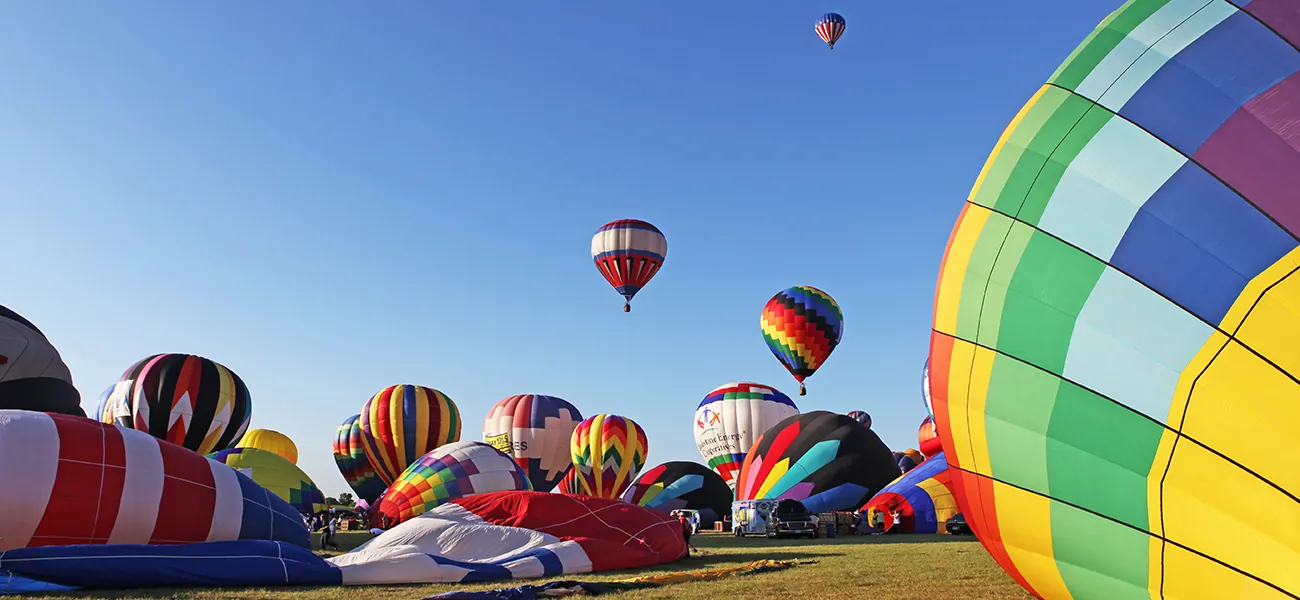 Una fotografía muestra una veintena de coloridos globos aerostáticos en distintas fases de inflado. Algunos están desinflados y otros están inflados. Tres de los globos no están en el suelo y son visibles contra un cielo azul brillante.