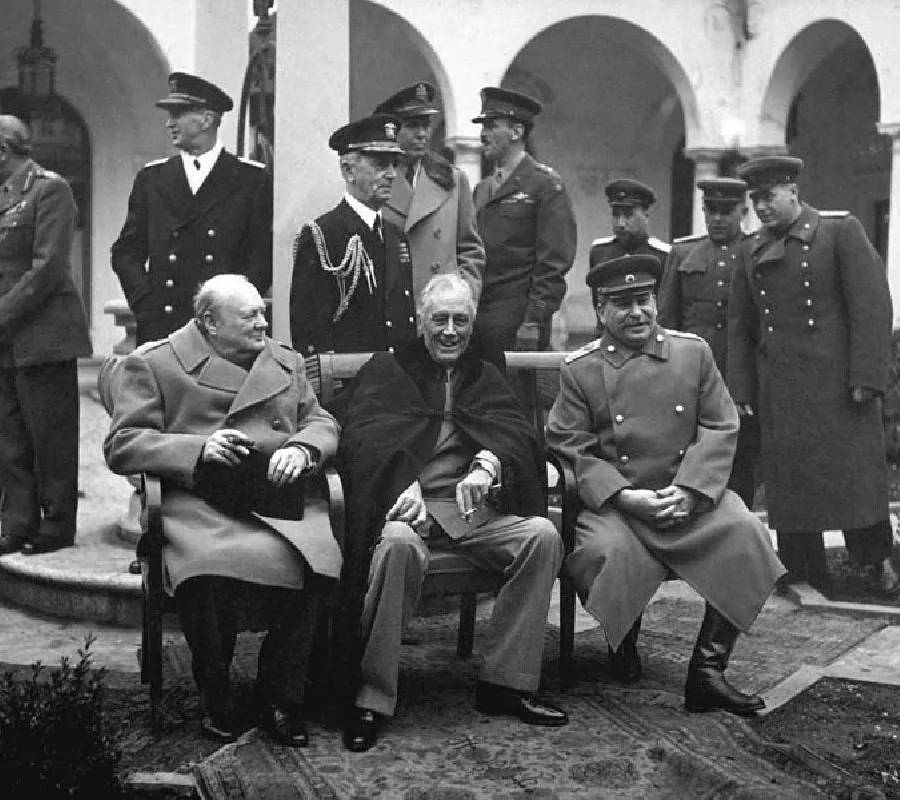 In this black and white photograph, three men sit in chairs next to each other. The man on the far left, is wearing a long winter coat and looking to his left. The man in the middle has a cape draped on his shoulders. He looks forward and smiles. The man on the right sits straight up and wears a military coat, tall boots, and a military hat. He looks forward and smiles. Behind the three men are eight white men in various military uniforms, facing different ways, and chatting with each other. They appear to be outside a building with arches. There are various ornate rugs placed on the floor under the chairs.