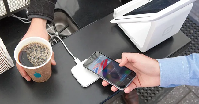A photo shows a point-of-sale system. A person is tapping their phone on a payment device while another person gives them a cup of coffee.