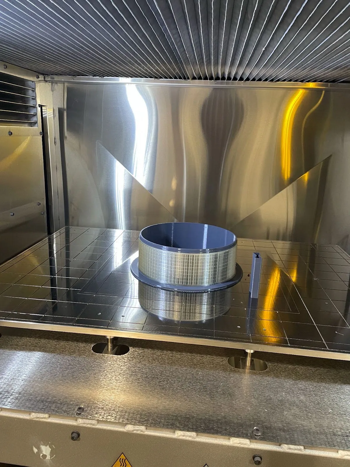  A metal cylinder sits on the printing bed in a metal 3d printer. The cylinder appears to be about forty centimeters in diameter. It’s outer surface is a textured, light metal. It’s inner surface is a solid color and appears to be polished smooth.