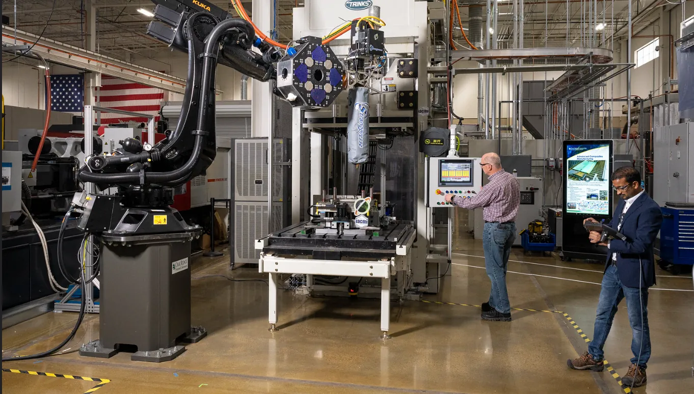 Two people stand near a large machine with a very large robotic arm that holds a large printing device. The arm is about three meters tall and about half a meter wide. The printing device hangs over a large metal work bed that stands in front of  the main body of another part of the machine. One of the people reviews an instrument panel and graphical display, while another holds what appears to be a tablet or similar device. 